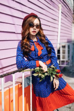 Mavi ve kırmızı retro elbiseli esmer kadın kırmızı gülleri tut ve dirseğini pembe duvardaki beyaz barikata koy..