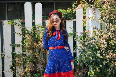 Mavi ve kırmızı retro elbiseli dalgalı esmer kadın beyaz çitin önünde duruyor, çalılarla, yatay.