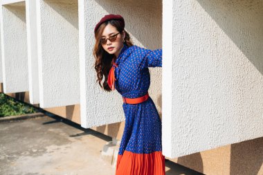 Mavi ve kırmızı retro elbiseli dalgalı esmer kadın binanın beyaz çerçevesinde duruyor ve vücudunu dışarıya doğru eğiyor..