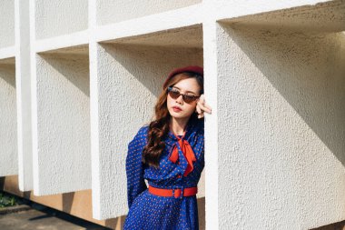 Mavi ve kırmızı retro elbiseli dalgalı esmer kadın binanın beyaz çerçevesinde duruyor ve başını çerçeveye dayıyor..