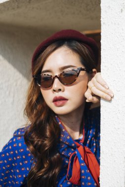 Mavi ve kırmızı retro elbiseli dalgalı esmer kadın binanın beyaz çerçevesinde duruyor ve kafasını çerçeveye dayayıp, kapatıyor..