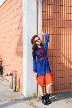 Mavi ve kırmızı retro elbiseli dalgalı esmer kadın güneş ışığında turuncu duvarın önünde duruyor ve beyaz direği tutuyor..