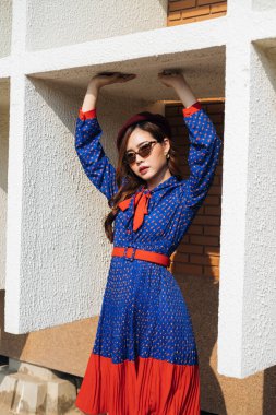 Mavi ve kırmızı retro elbiseli dalgalı esmer kadın binanın beyaz çerçevesinde duruyor ve üst kısma dokunuyor..