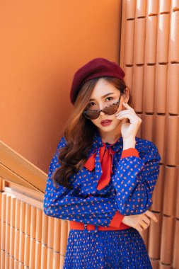 Mavi ve kırmızı retro elbiseli dalgalı esmer kadın turuncu duvarda egzoz dumanıyla dikiliyor ve kameraya bakın..