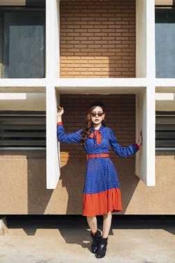 Mavi ve kırmızı retro elbiseli dalgalı esmer kadın resmin ortasında, binanın beyaz çerçevesinde duruyor..