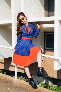 Dalgalı esmer, mavi ve kırmızı retro elbiseli kadın yamaçtaki beyaz tırabzanda oturuyor..