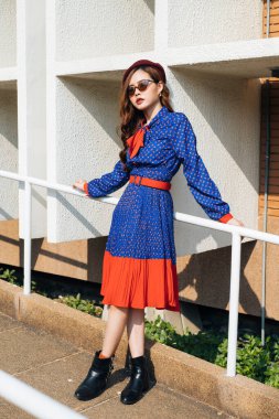 Dalgalı esmer, mavi ve kırmızı retro elbiseli kadın yamaçtaki beyaz tırabzanlara yaslanıyor..