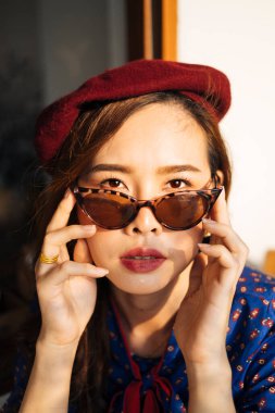 Mavi ve kırmızı retro elbiseli ve güneş gözlüklü esmer kadın cam pencerenin yanındaki tahta masada baykuşla oturuyor..