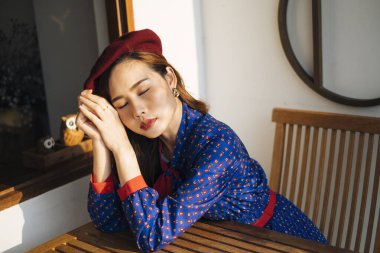 Mavi ve kırmızı retro elbiseli esmer kadın cam pencerenin yanındaki tahta masada baykuşla oturuyor..