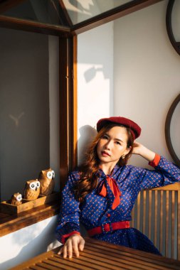 Mavi ve kırmızı retro elbiseli dalgalı esmer kadın güneş ışığında baykuşlu cam pencerenin yanındaki ahşap masada oturuyor..