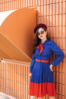 Dalgalı esmer, mavi ve kırmızı retro elbiseli kadın turuncu duvarın önünde kendine güvenen egzoz dumanıyla duruyor..