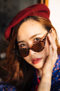 Mavi ve kırmızı retro elbiseli, güneş gözlüklü, camlı pencerenin yanındaki tahta masada oturan ve başını eğen dalgalı esmer kadın..