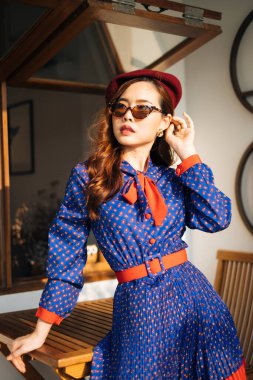 Dalgalı esmer kadın, mavi ve kırmızı retro elbiseli ve güneş gözlüklü, gün ışığında ahşap masaya yaslanmış..