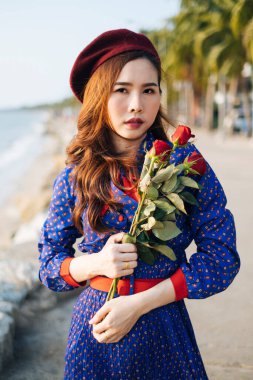 Mavi ve kırmızı retro elbiseli dalgalı esmer kadın sahilde duruyor ve güllere sarılıyor..