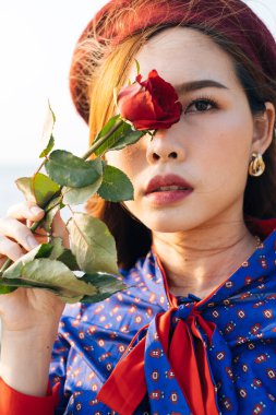 Dalgalı esmer, mavi ve kırmızı retro elbiseli kadının yakın çekimi. Sağ gözünü gülle gizliyor..