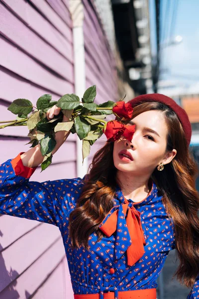 波ブルネット女性で青と赤のレトロドレスホールド赤いバラで彼女の顔と立ってでザピンクレンガと木の壁で太陽の光 — ストック写真