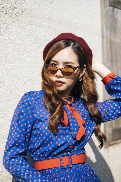 Mavi ve kırmızı retro elbiseli esmer kadının orta boy portresi. Eski evin duvarında güneş ışığında serin bir duruş sergiliyor..