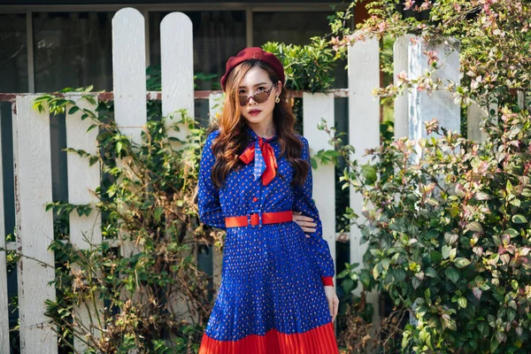 Mavi ve kırmızı retro elbiseli dalgalı esmer kadın beyaz çitin önünde duruyor, çalılarla, yatay.