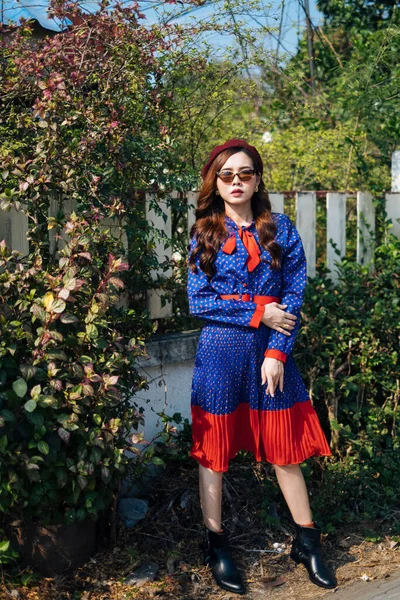 Dalgalı esmer, mavi ve kırmızı retro elbiseli kadın beyaz çitin orada çalılarla dikiliyor..