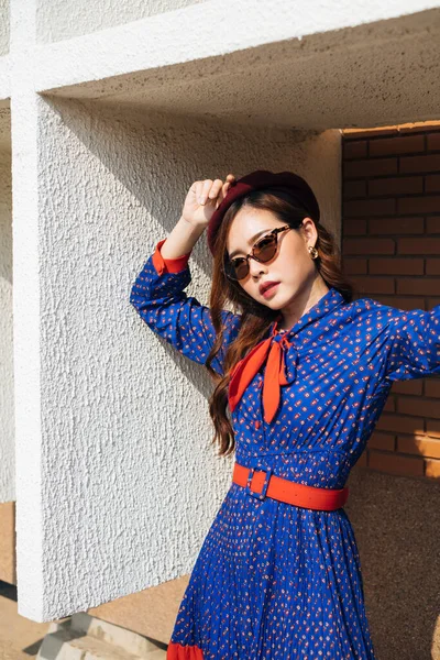 Dalgalı esmer, mavi ve kırmızı retro elbiseli kadın güneş ışığında binanın beyaz çerçevesinde duruyor..