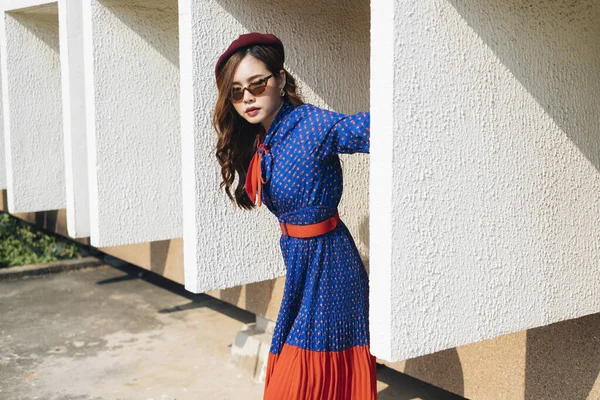 Mavi ve kırmızı retro elbiseli dalgalı esmer kadın binanın beyaz çerçevesinde duruyor ve vücudunu dışarıya doğru eğiyor..