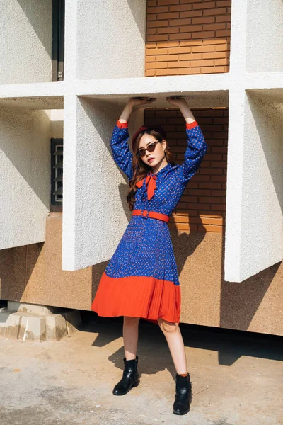 Mavi ve kırmızı retro elbiseli dalgalı esmer kadın binanın beyaz çerçevesinde duruyor ve üst kısma dokunuyor..