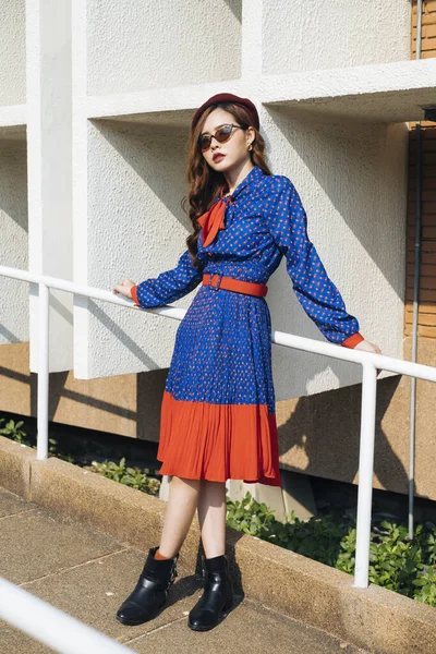 Dalgalı esmer, mavi ve kırmızı retro elbiseli kadın yamaçtaki beyaz tırabzanlara yaslanıyor..