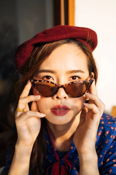 Mavi ve kırmızı retro elbiseli ve güneş gözlüklü esmer kadın cam pencerenin yanındaki tahta masada baykuşla oturuyor..