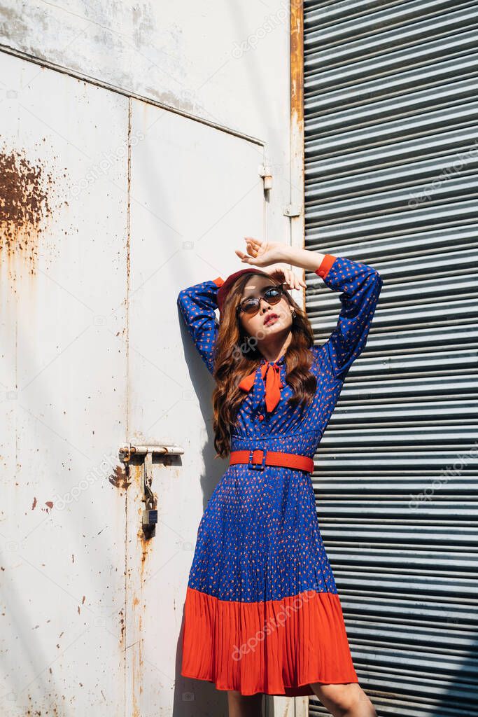Wavy brunette woman in blue and red retro dress and sunglasses standing like a supermodel at the old warehouse with metal shutter door.