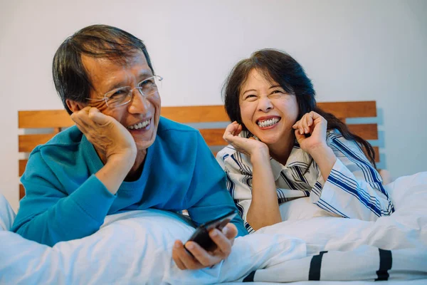 ベッドルームで一緒にテレビを見て陽気なアジアの高齢者のカップル 彼らはパネルを制御するためにスマートフォンを使って — ストック写真