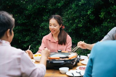 Genç Asyalı kadın yemek çubuğu kullanıyor. Bahçede ailesiyle barbekü yiyor..