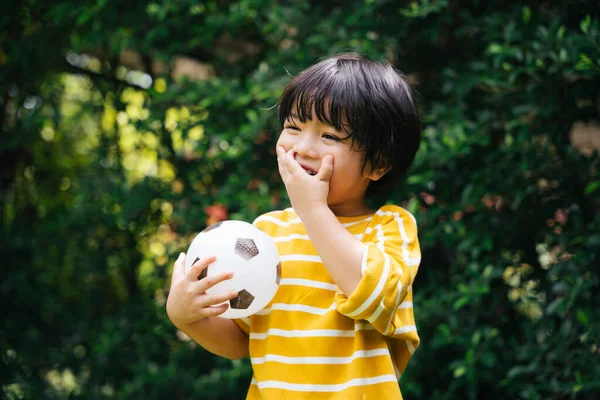 Cheerful asian kid holding a football cover mouth with hand laughing and smile at park.