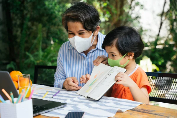 Grandmother teaching grandson how to drawn in a book. Senior elderly old and son learn drawing online with laptop. Both wear mask.