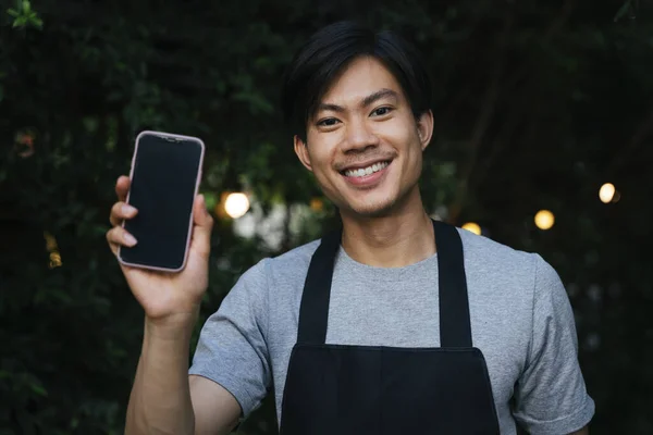 Cheerful asian waiter wear black apron showing blank screen mock up smartphone for copy space