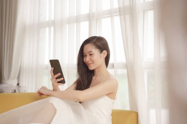 Beyaz elbiseli Asyalı genç bir kadın oturma odasındaki koltukta akıllı telefon kullanıyor..