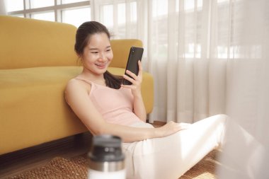 Genç Asyalı güzel kadın evde komik ve mutlu bir ifadeyle akıllı telefon kullanıyor..