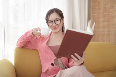 Pembe ceketli, gözlüklü, kitap okuyan ve kahve içen Asyalı genç bir kadın..