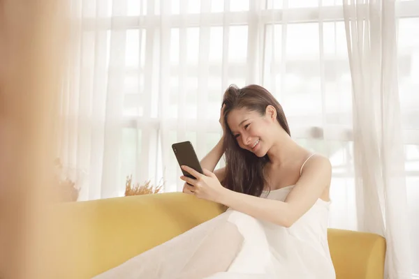 若いですアジアの美しさの女性で白いドレス使用してスマートフォン上のAソファでリビングルーム — ストック写真