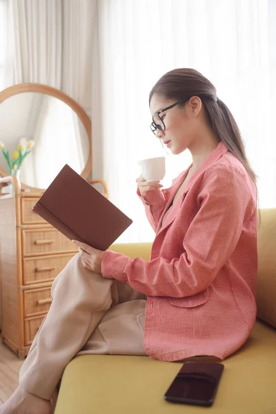 Yan görünüş - Genç Asyalı güzel kadın ceket giyiyor ve gözlük takıyor kahve içiyor ve kanepede kitap okuyor.