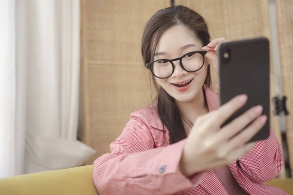 若いアジアの美しさの女性は眼鏡を着用し ピンクのジャケットライブストリームとともにスマートフォン — ストック写真