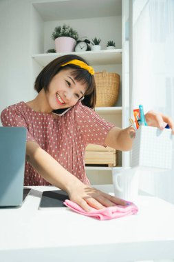 Asya Tayland Üniversitesi öğrencisi kuru bezle masayı temizliyor ve oturma odasında telefonla konuşuyor..