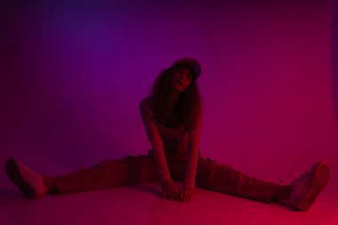 Genç Asyalı kadın neon ışıklı arka planda yerde oturan şapka ve süveter giyiyor..