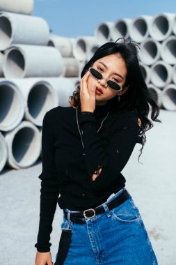 Siyah kazaklı ve kot pantolonlu moda kızı büyük beton borularla dolu sessiz bir yerde duruyor..