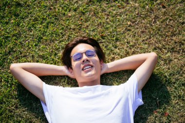 Beyaz tişörtlü, gözlüklü, siyah saçlı adamın güneş ışığında sahada uzandığı orta boy yakın çekim..