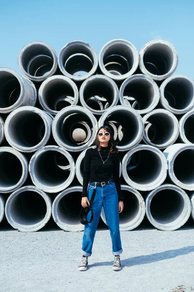 Fashionista girl in black sweater and jeans standing in front of big concrete tube in summer season.