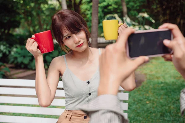 アジア系タイ人のカップルがスマートフォンで写真を撮る 女性は ベンチに座って2つの赤と黄色のカップの屋外を保持 スマートフォンで写真を撮る男 — ストック写真