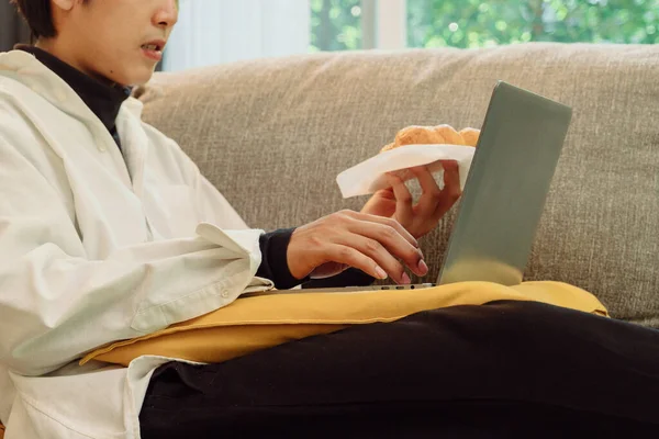 商人在家里工作 一边吃新鲜的烤羊角面包一边用笔记本电脑 — 图库照片