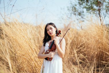 Beyaz elbiseli siyah, uzun saçlı kemancı kadın otlakta kemanıyla duruyor..