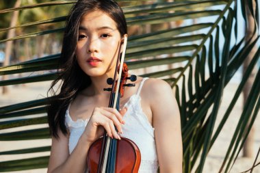 Beyaz elbiseli ve keman çalan siyah uzun saçlı kemancı kadın hindistan cevizi yaprağının önünde duruyor..