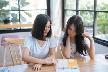 Kafede oturan iki beyaz tişörtlü kız içeceği seçmek için menüye bakıyorlar..